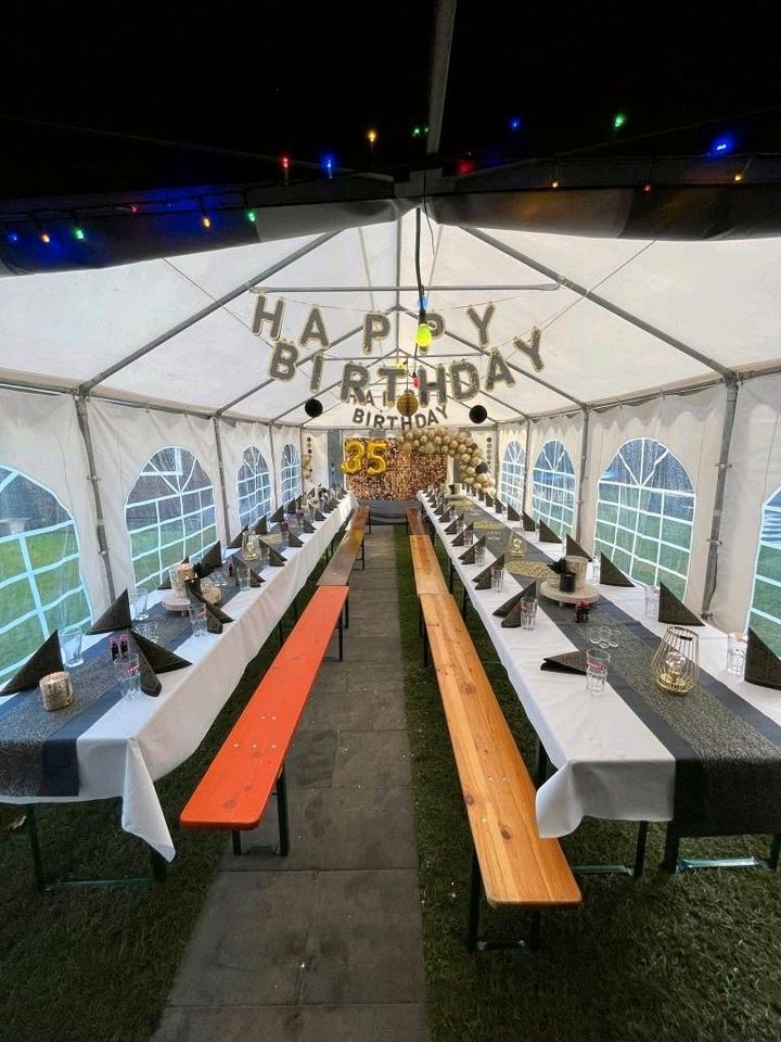 Ein für eine Geburtstagsfeier aufgebautes Zelt mit zwei langen, mit weißen Tischdecken und dunklen Tischläufern bedeckten Tischen, bereit für die Gäste.
