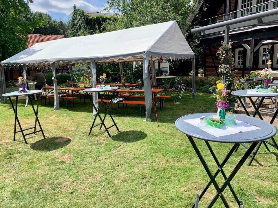Eine Terrasse mit weißem Pavillon, Picknickbänken und mehreren runden Stehtischen auf einer Rasenfläche.