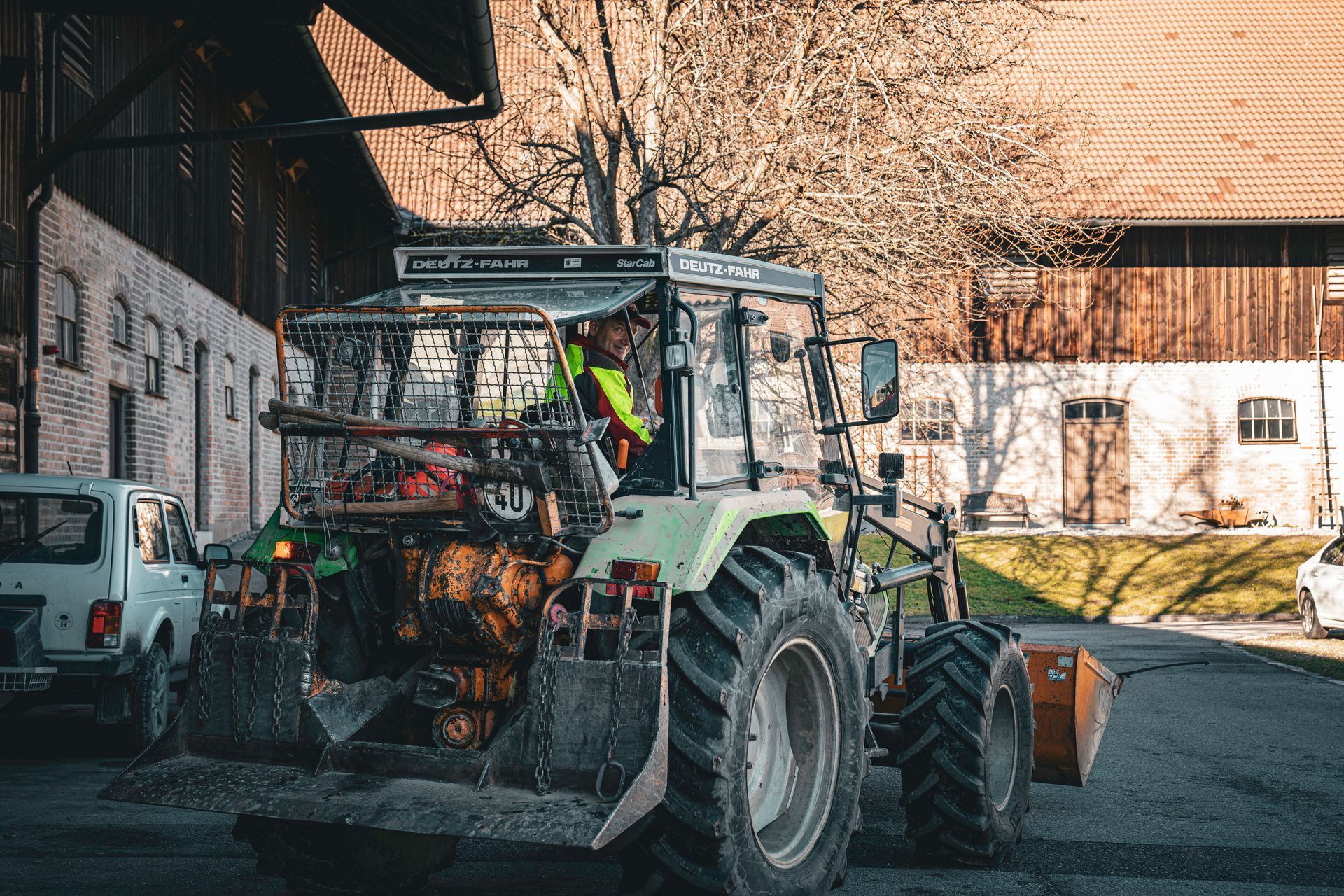 Ein Traktor mit Frontlader, Heuballen und einer Scheune im Hintergrund steht auf einem Bauernhofhof.