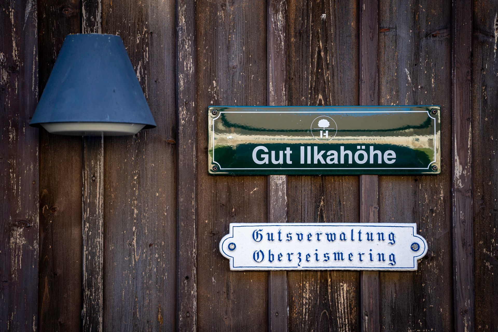 Ein grün-weißes Schild mit der Aufschrift „Gut Ilkahöhe“ an einer verwitterten Holzwand, darüber eine blaue Wandlampe und darunter ein weiteres Schild.