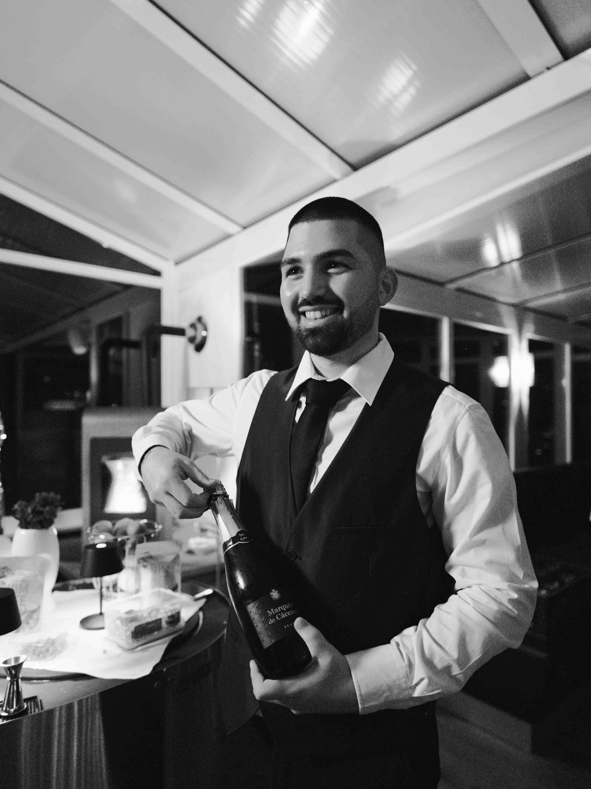 lachender Barkeeper der einen hochwertigen Sekt Korken platzen lässt