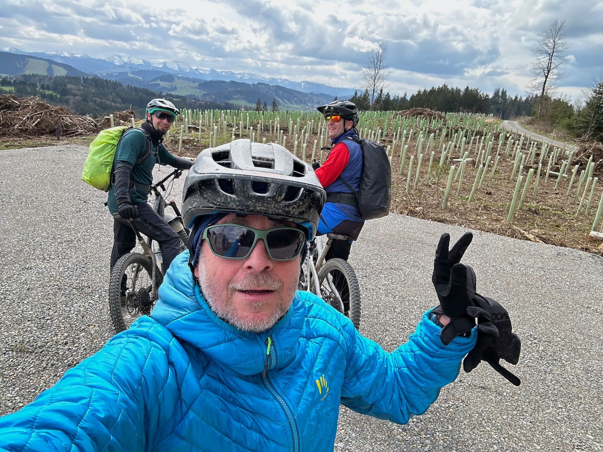 Drei Radfahrer auf Fahrrädern in einem Schottergebiet mit Bergen im Hintergrund, einer macht ein Selfie.