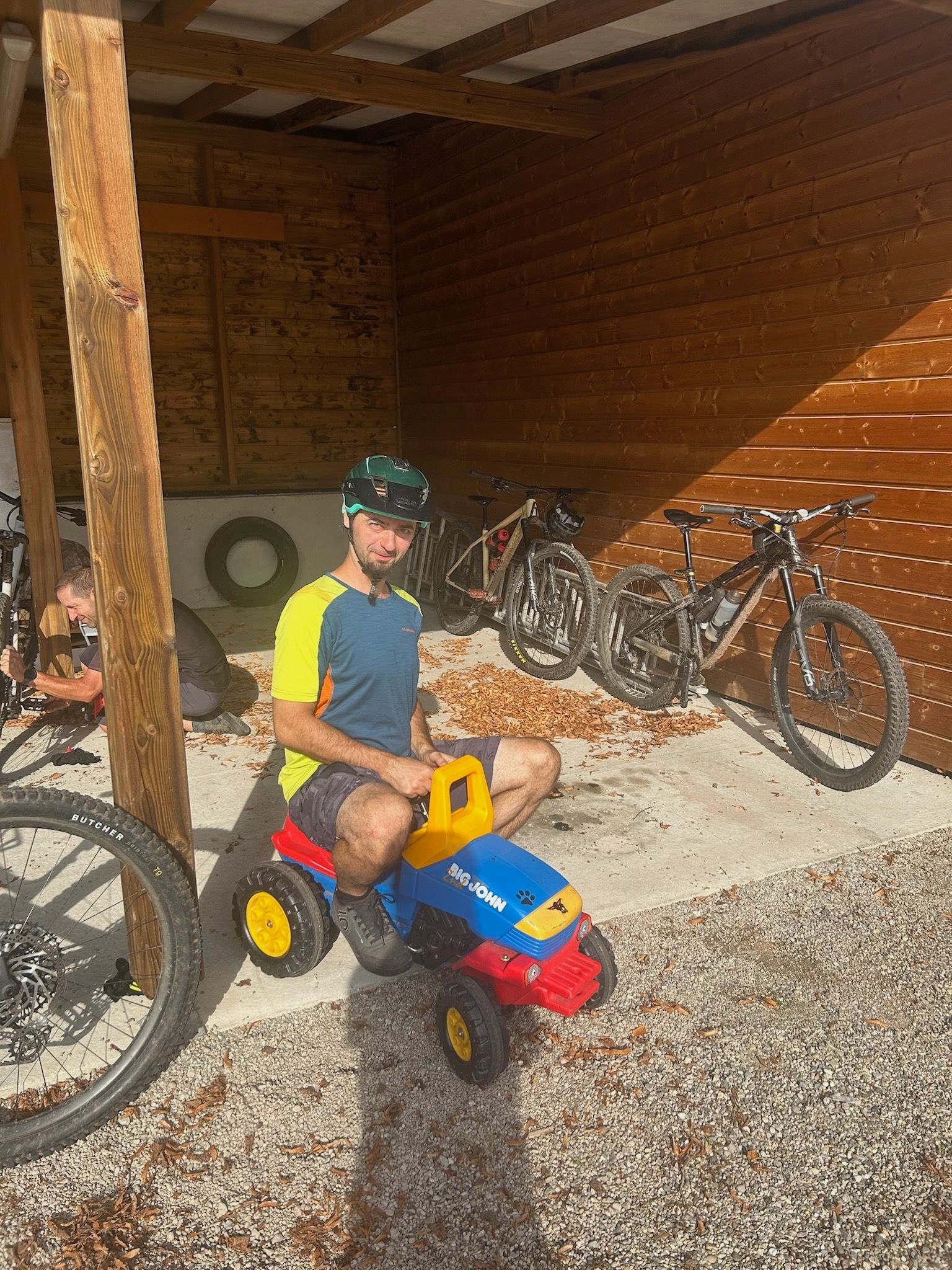 Ein Mann sitzt im Freien auf einem Spielzeugtraktor; im Hintergrund sind Mountainbikes zu sehen.