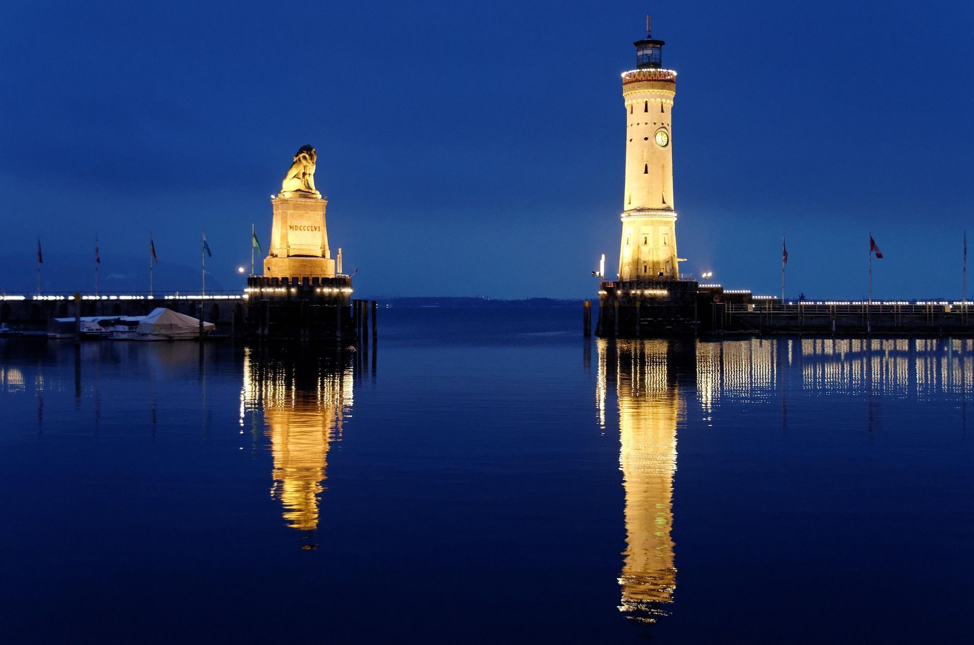 Beleuchteter Leuchtturm und Löwenstatue in der Abenddämmerung, die sich im dunkelblauen Wasser spiegeln.