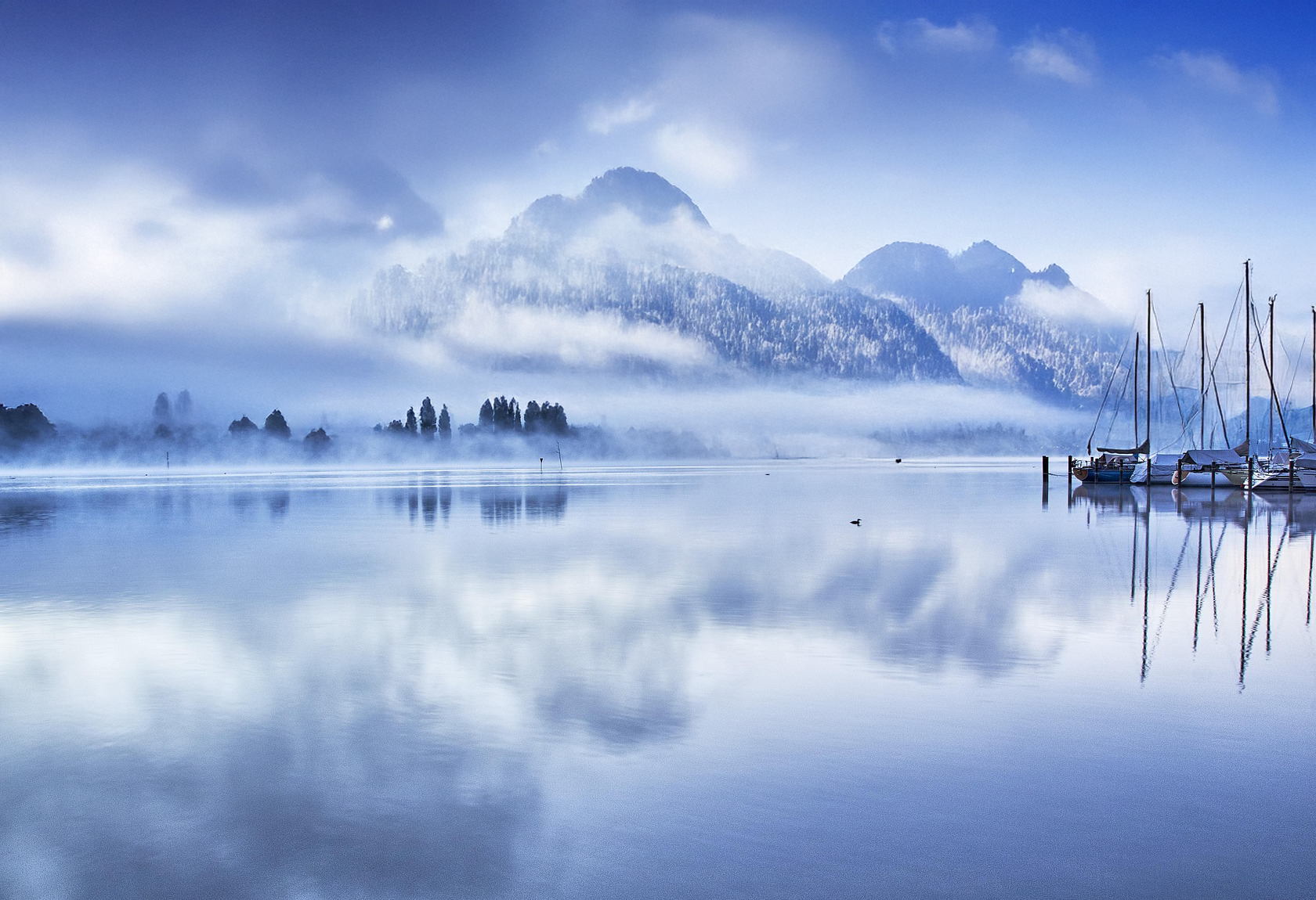 Nebliger See mit Spiegelungen. Segelboote liegen vor Anker. Neblig, blau und friedlich.