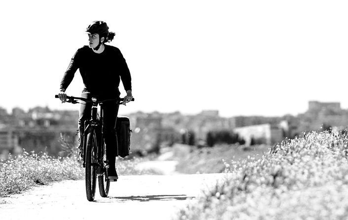 Eine Person fährt mit dem Fahrrad auf einem unbefestigten Weg. Sie trägt einen Helm und schwarze Kleidung. Im Hintergrund sind Gebäude zu sehen.