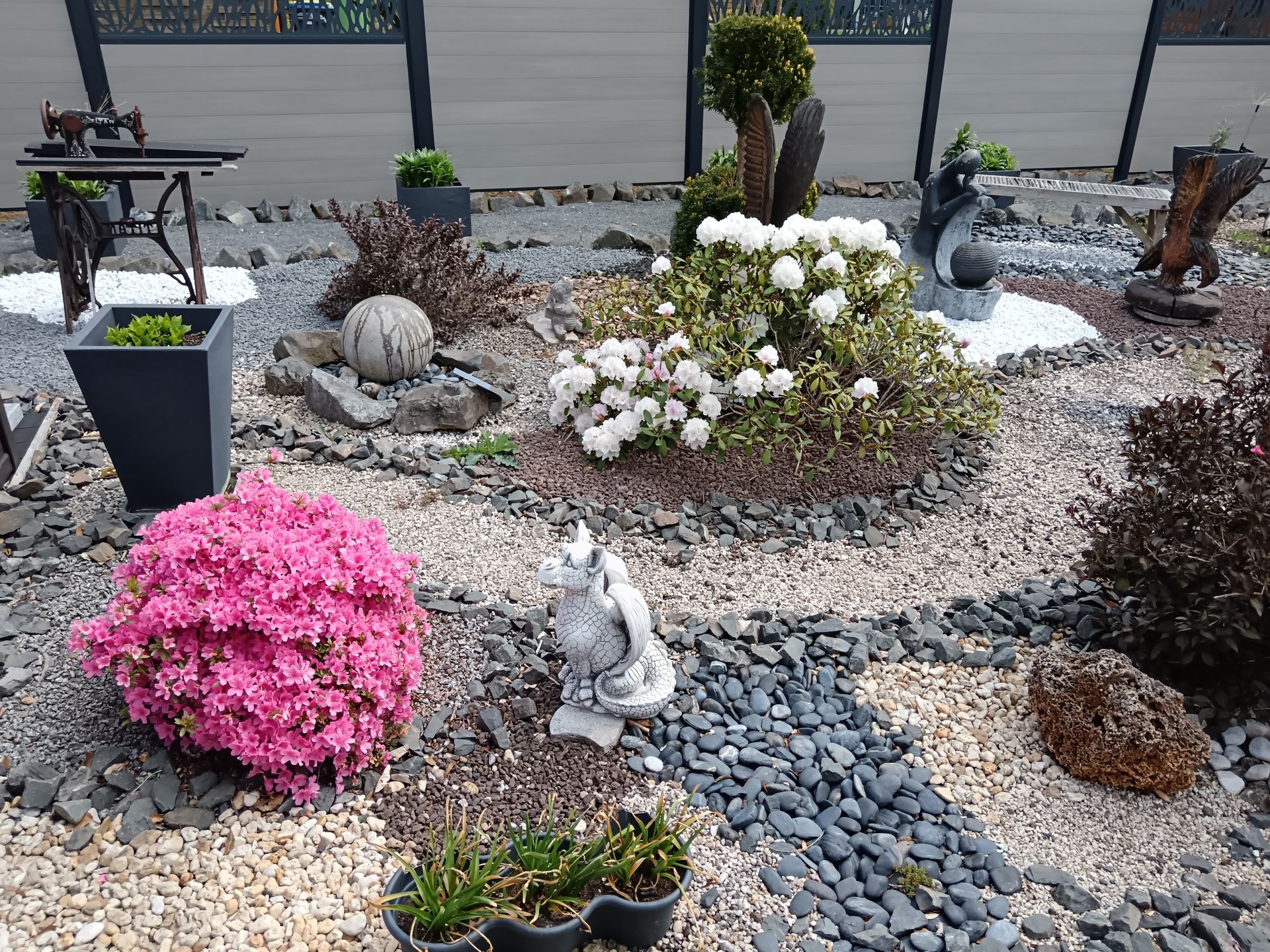 Ein Steingarten mit einer Vielzahl von Pflanzen, darunter ein leuchtend rosa blühender Strauch und eine Steinskulptur.