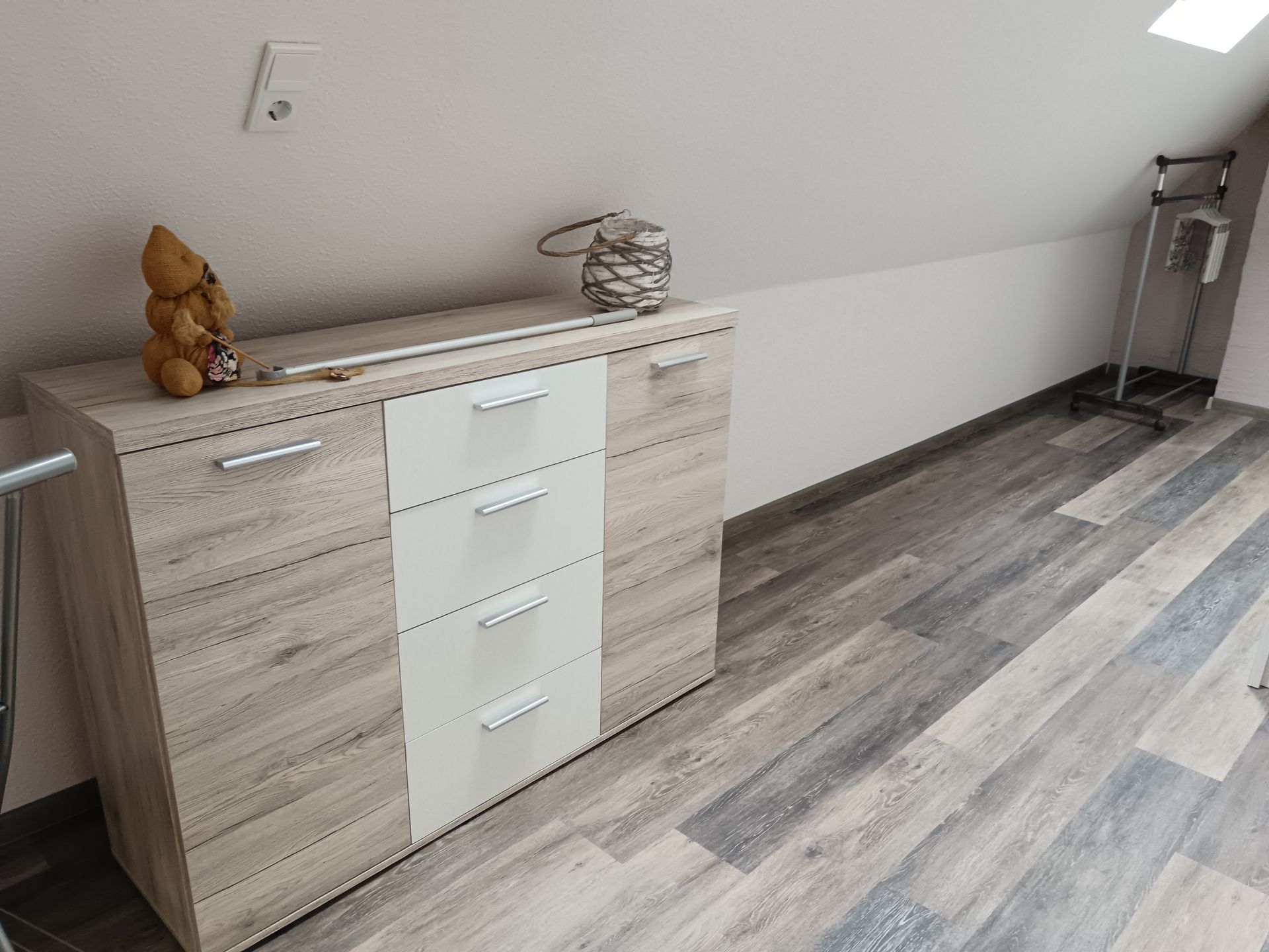 Holzkommode mit Schubladen und Dekorationsgegenständen an einer schrägen Wand mit Holzboden.