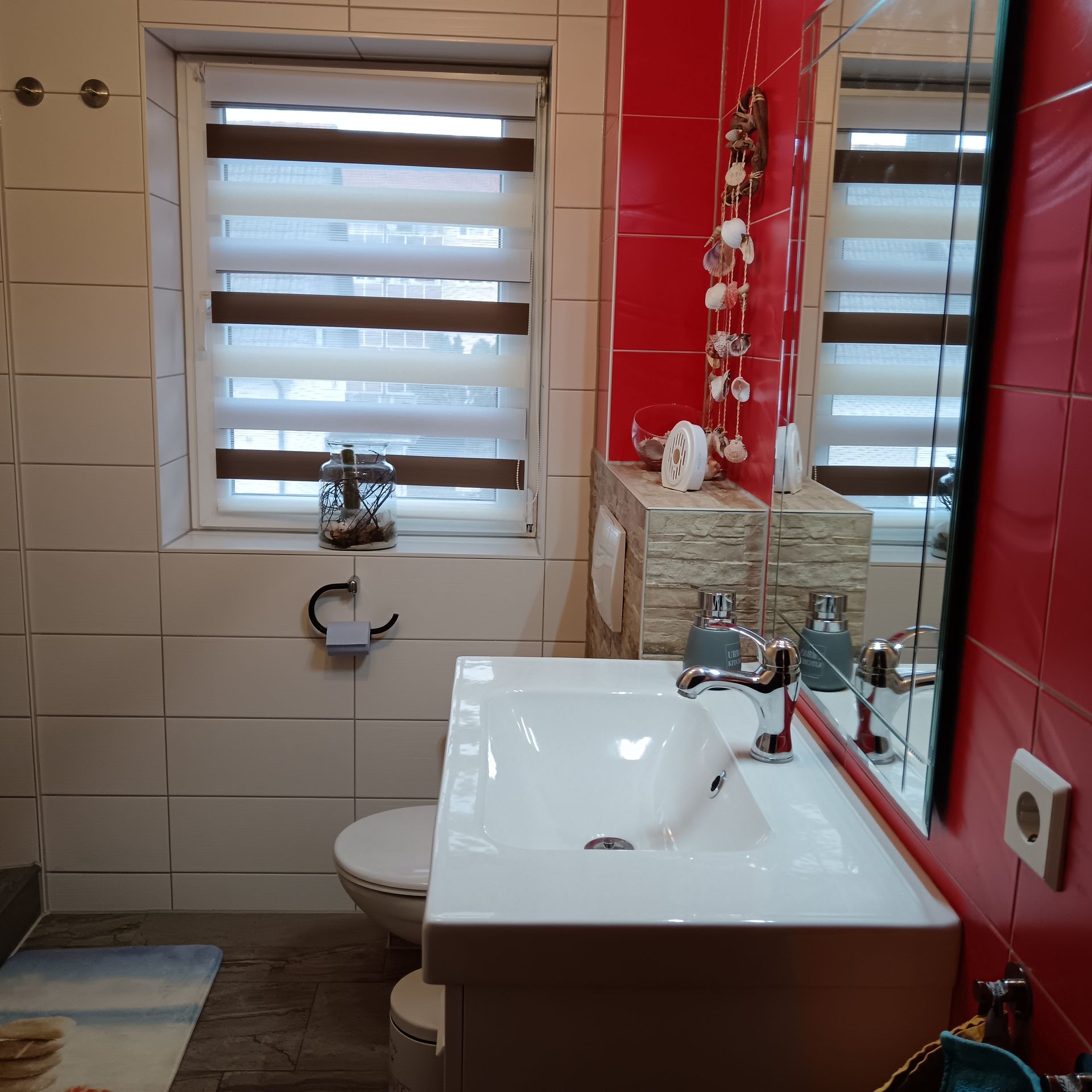 Badezimmer mit weißem Waschbecken, Toilette, roter Wand, Fenster und dekorativen Muscheln.