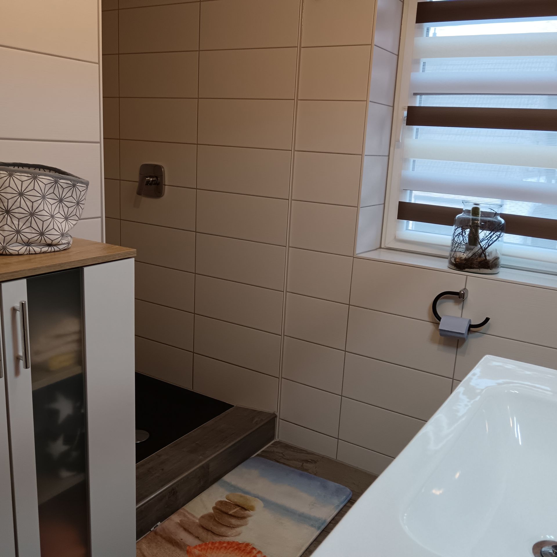 Badezimmer mit Dusche, Waschbecken und Unterschrank. Beige geflieste Wände, Fenster mit Jalousien.