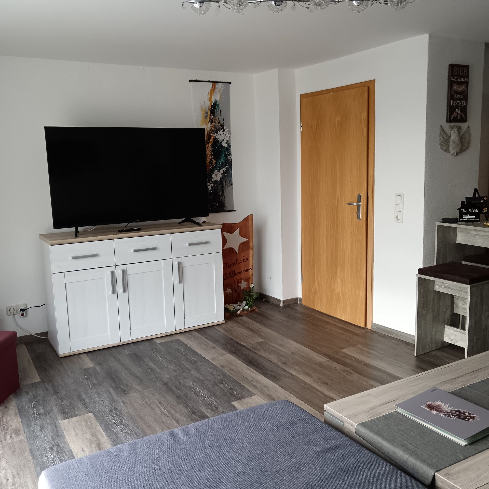 Wohnzimmer mit Fernseher auf weißem Schrank, Holztür und grauem Laminatboden.
