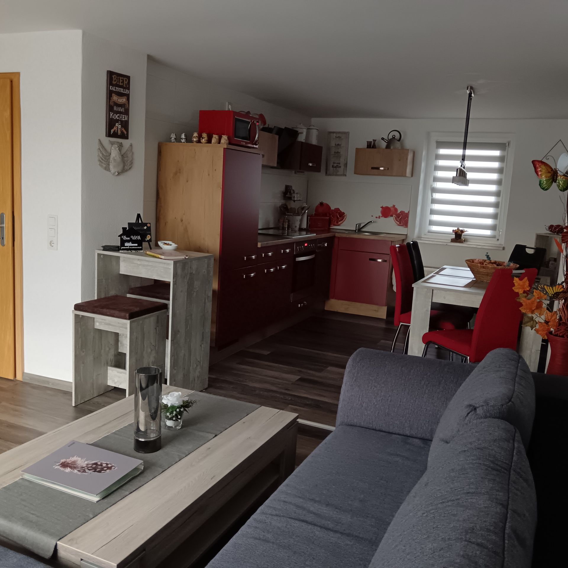 Wohnzimmer mit Küche im Hintergrund; graue Couch, Holzmöbel, rote Akzente, Fenster mit Jalousien.