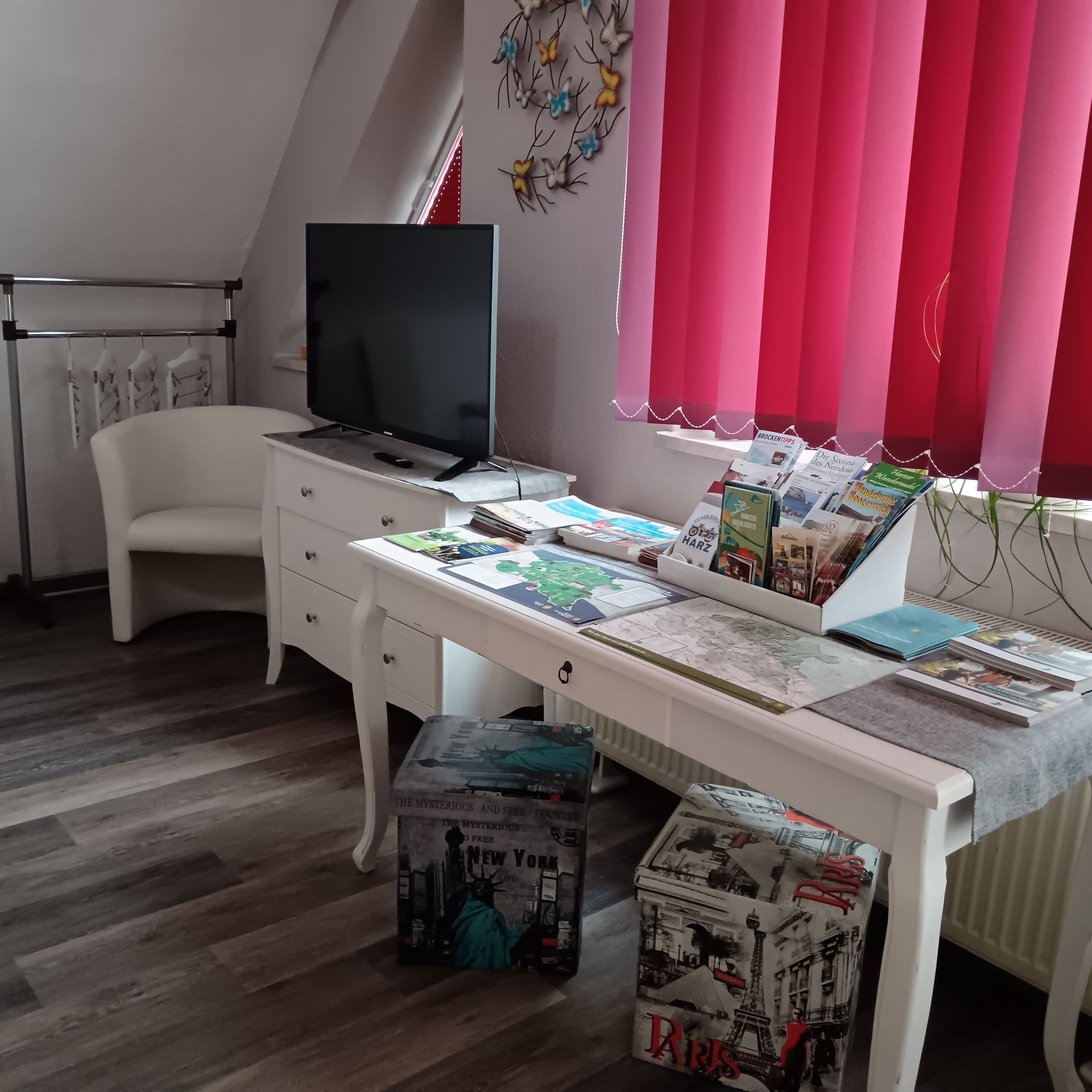 Weißer Schreibtisch mit Informationsmaterial, Fernseher, Stuhl, Kleiderständer und Fenstern mit roten Jalousien.