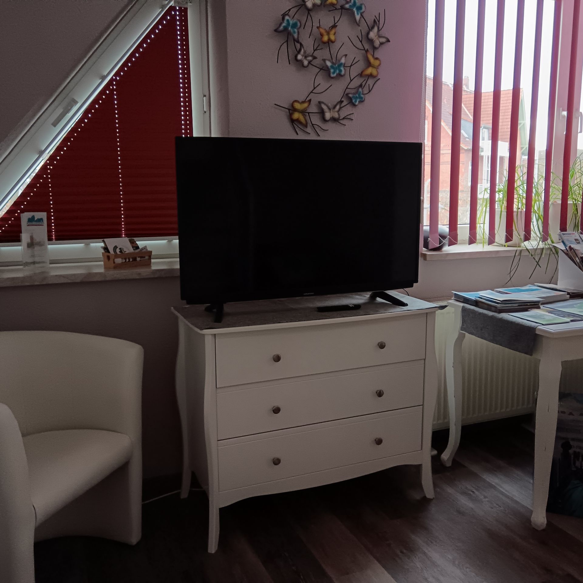 In einem Zimmer mit Fenster und Stühlen steht ein Fernseher auf einer weißen Kommode.