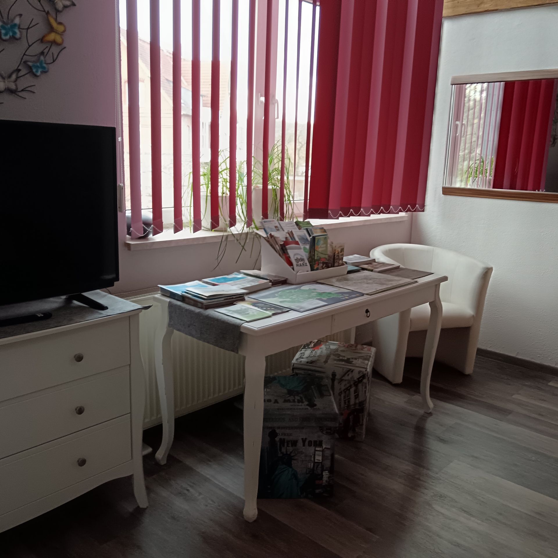 Weißer Schreibtisch mit Broschüren und Stuhl am Fenster mit roten Jalousien und Spiegel. Fernseher auf weißer Kommode.