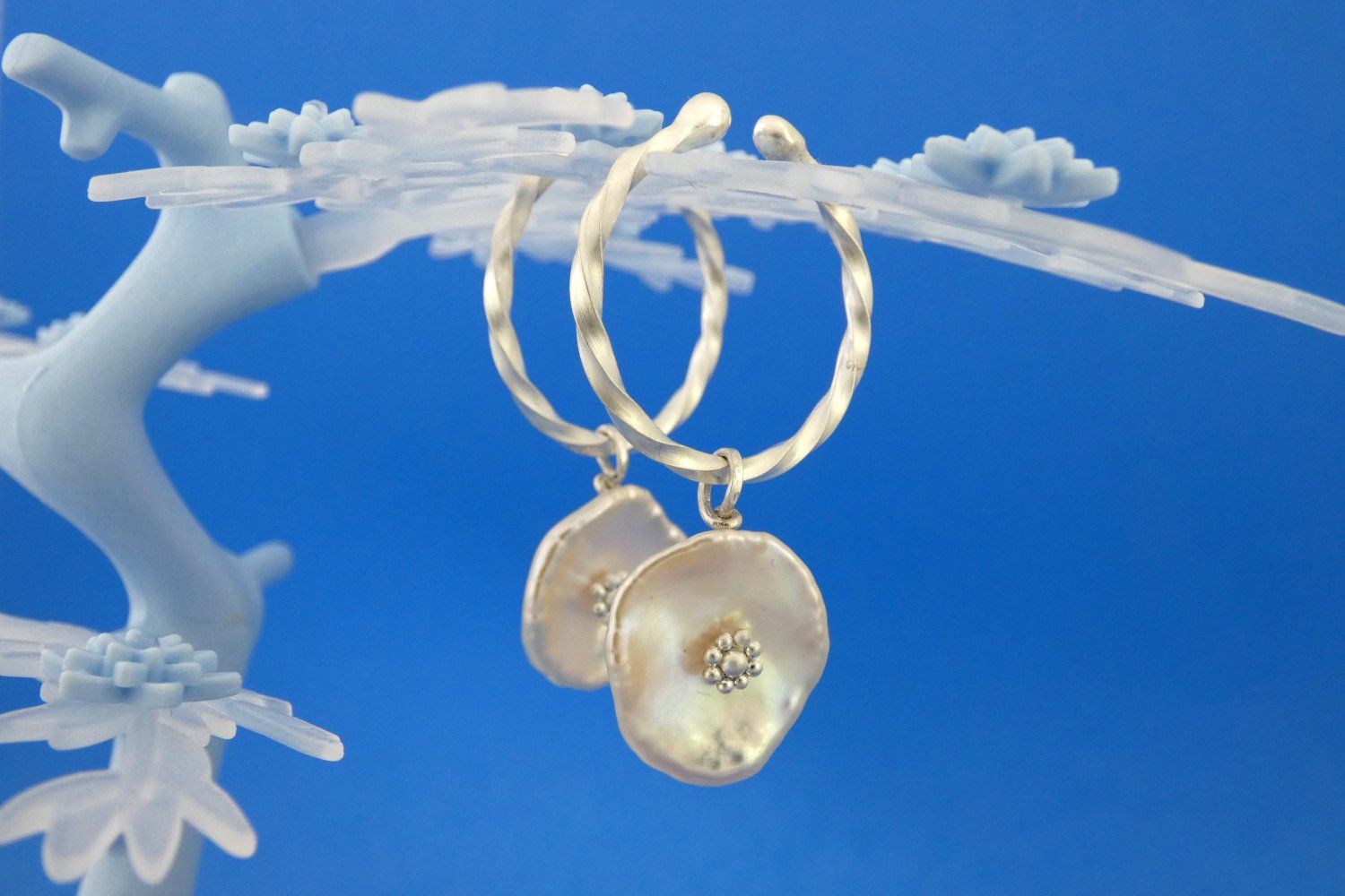 Creolen, die keiner Ohrlöcher bedürfen, gefertigt aus gedrehtem Vierkantdraht aus Argentium-Sterlingsilber, mit Anhängern aus großen flachen Keshi-Süßwasserperlen, zart rosafarben und mit stilisierten Silberblüten in der Mitte