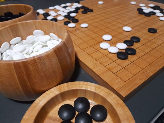 Neben zwei Bambusschalen mit weiteren Spielsteinen steht ein hölzernes Go-Brett mit schwarzen und weißen Steinen.