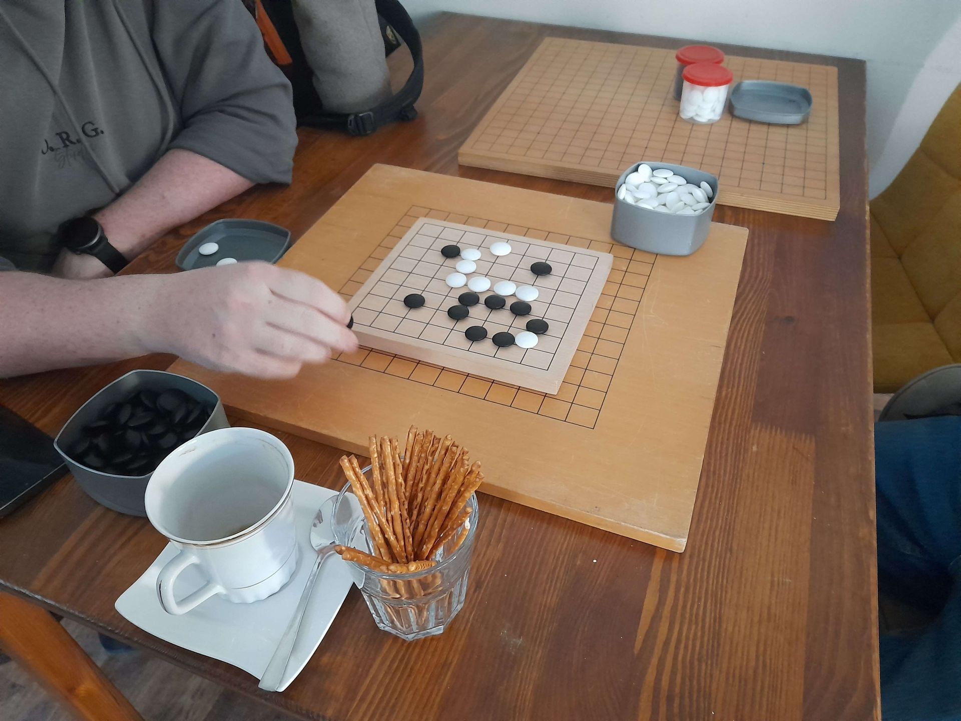 A person playing Go, a board game, with black and white stones on a wooden board at a table. Snacks and drinks are nearby.
