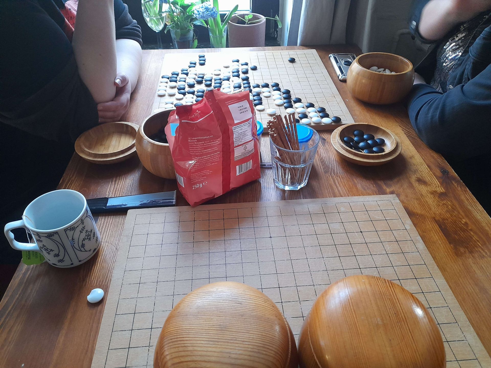 People playing Go, a board game, with black and white stones on a wooden table. Bowls, a drink carton, and snacks are also present.