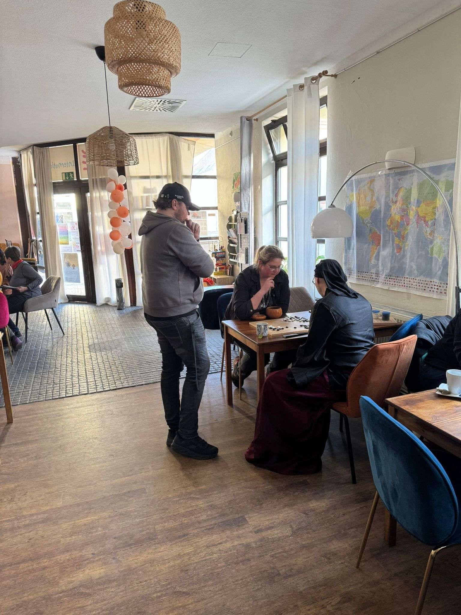 People at tables in cafe, some playing a board game. A man stands nearby. Interior with light wood floors.