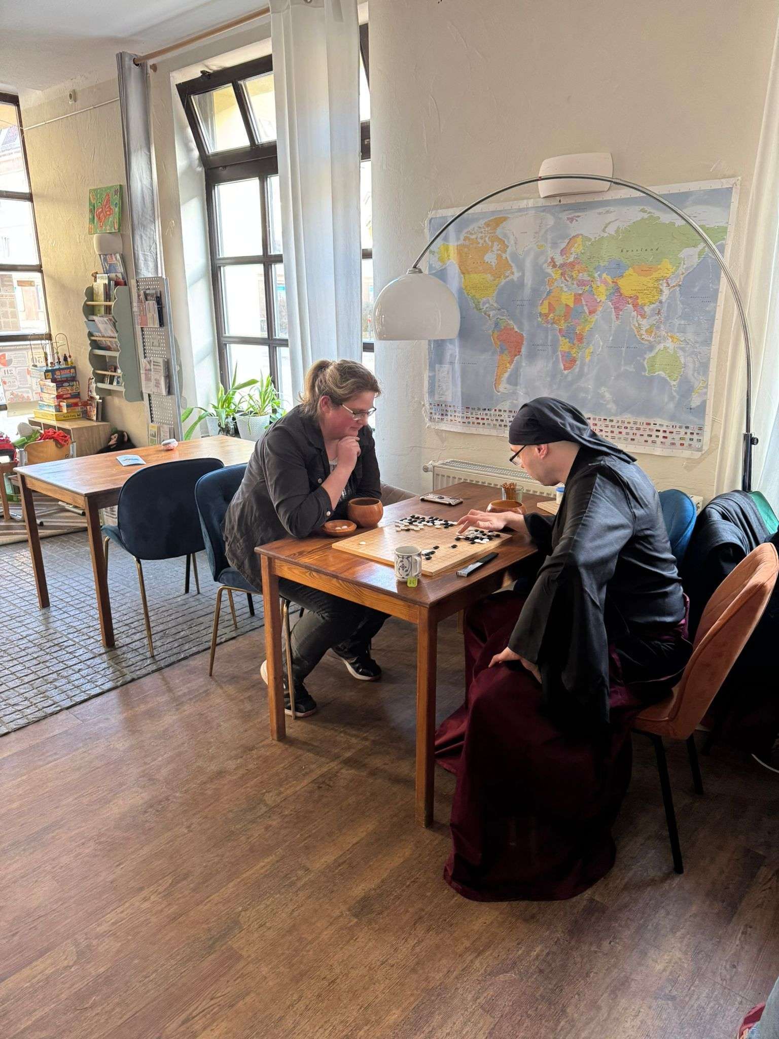 Two people playing a board game at a table in a room with a map and windows.