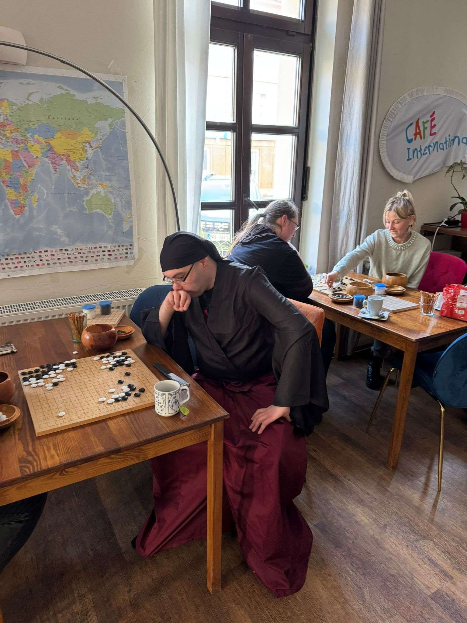 People playing Go at a cafe. Man in black robe ponders moves, others at tables.