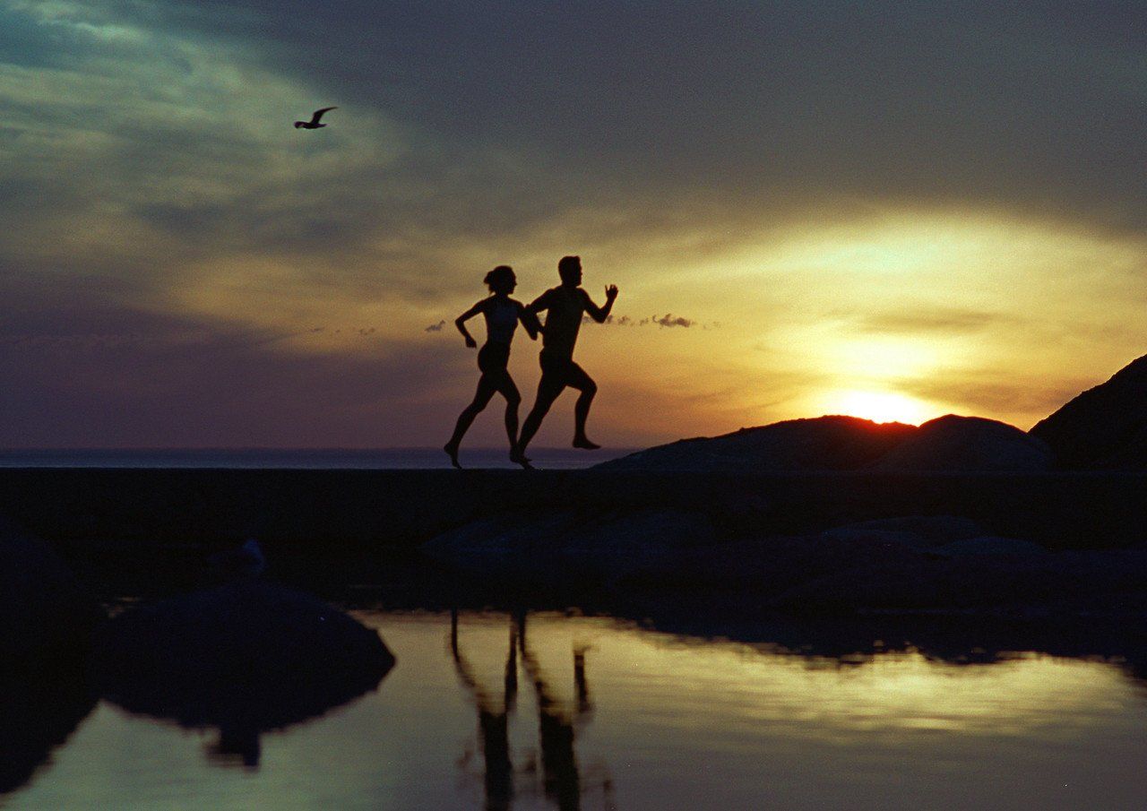 Silhouette of man and woman running close to water, with the sun setting in the background.