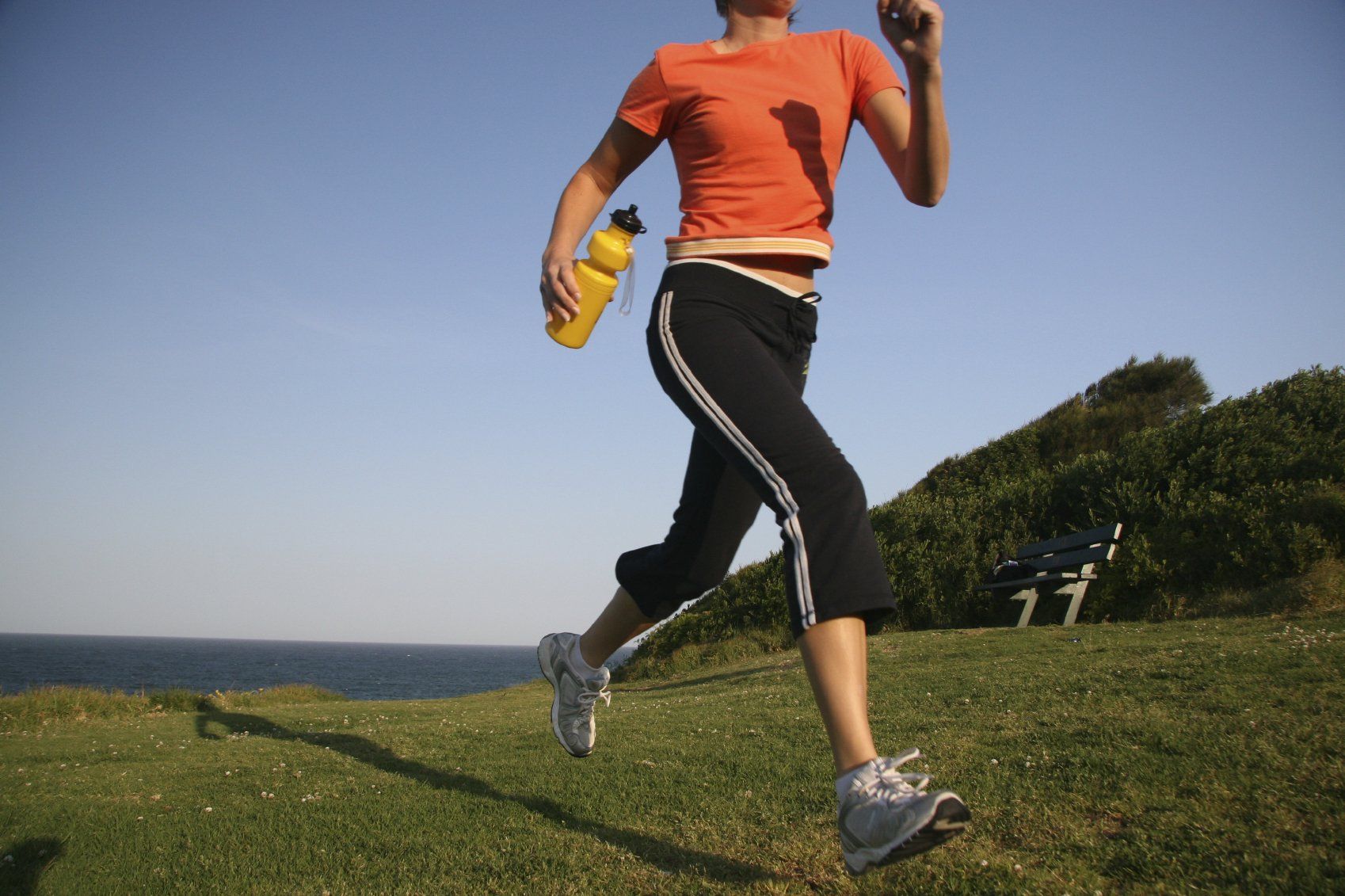 Young lady jogging across a grass field holding a yellow water bottle in her right hand.