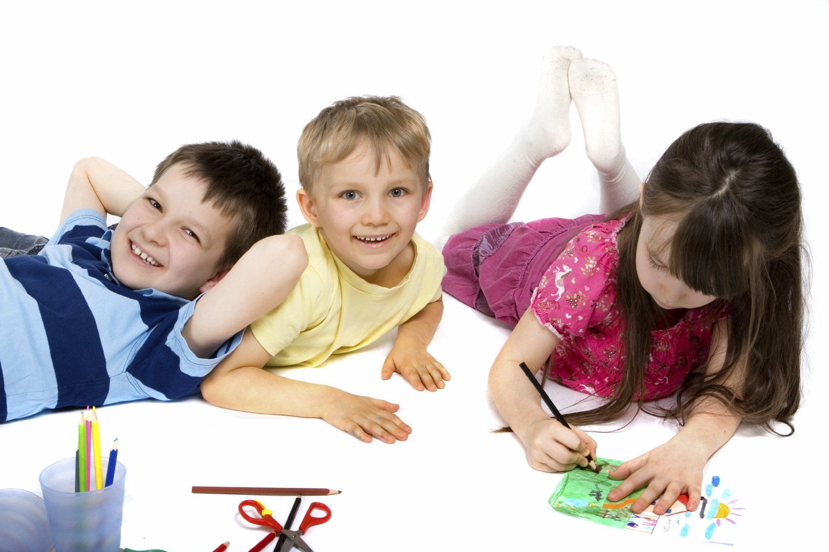 Two young boys and a young girl, in colorful clothing, lying on the flow and playing with crayons.