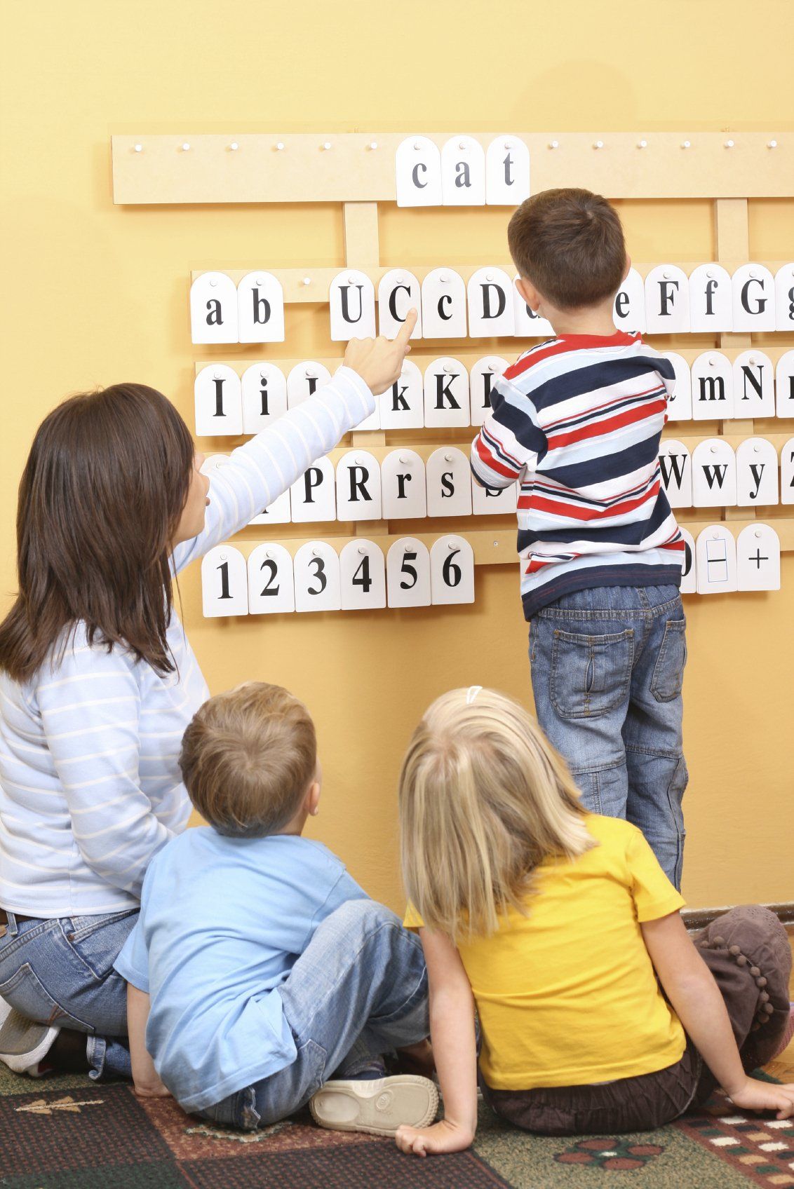 Teacher and two little kits sitting on floor while third kid arranges letters and numbers on  a wall rck.