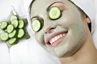 Frau mit Gurkenscheiben auf den Augen und einer grünen Gesichtsmaske, die während einer Spa-Behandlung lächelt.