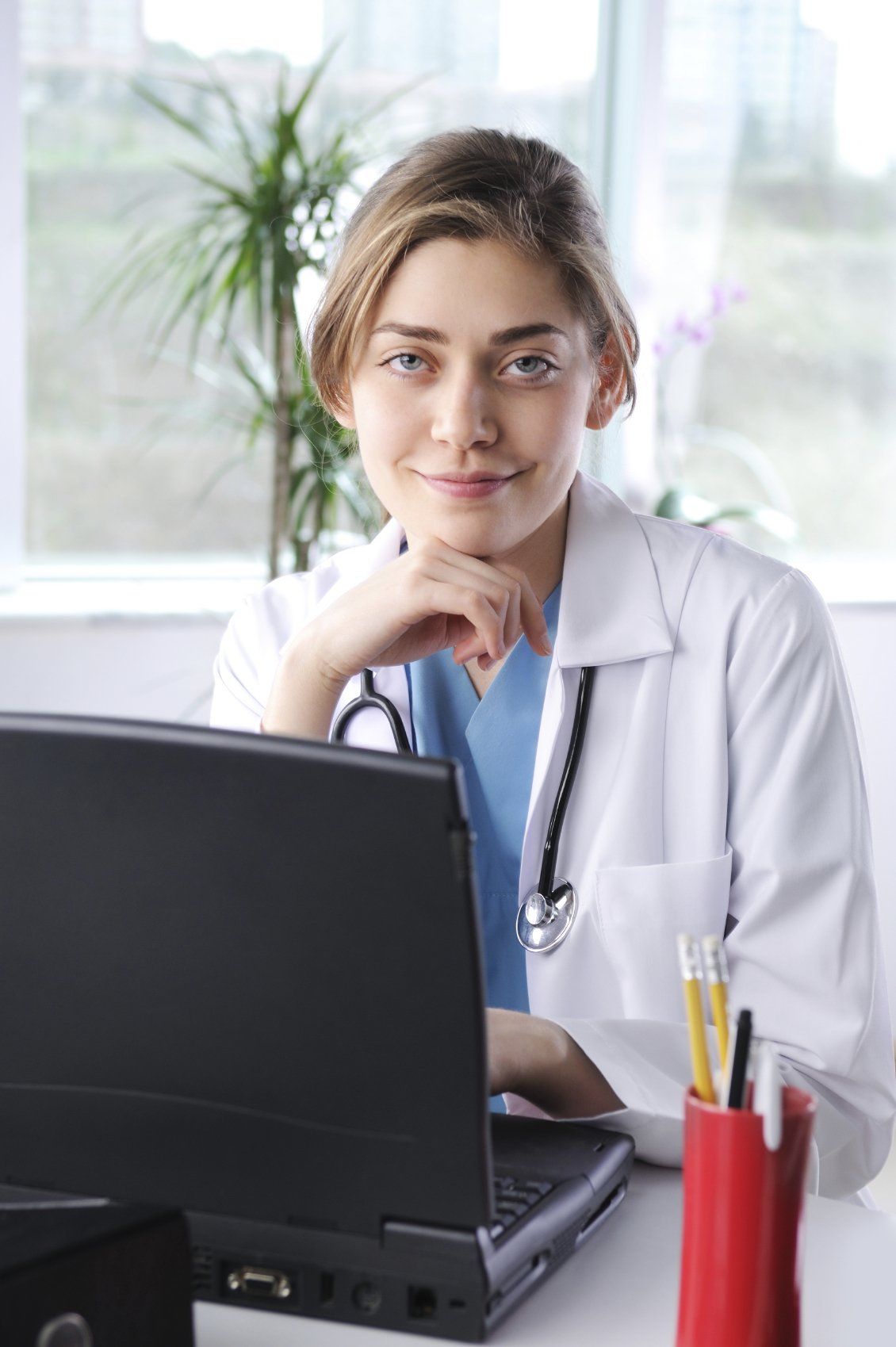 A young, female doctor in a white robe sitting in front of her black laptop.