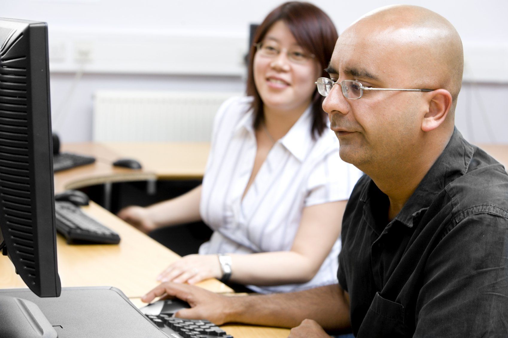 Bald man in black shirt sitting at computer with a smiling, Asian woman.