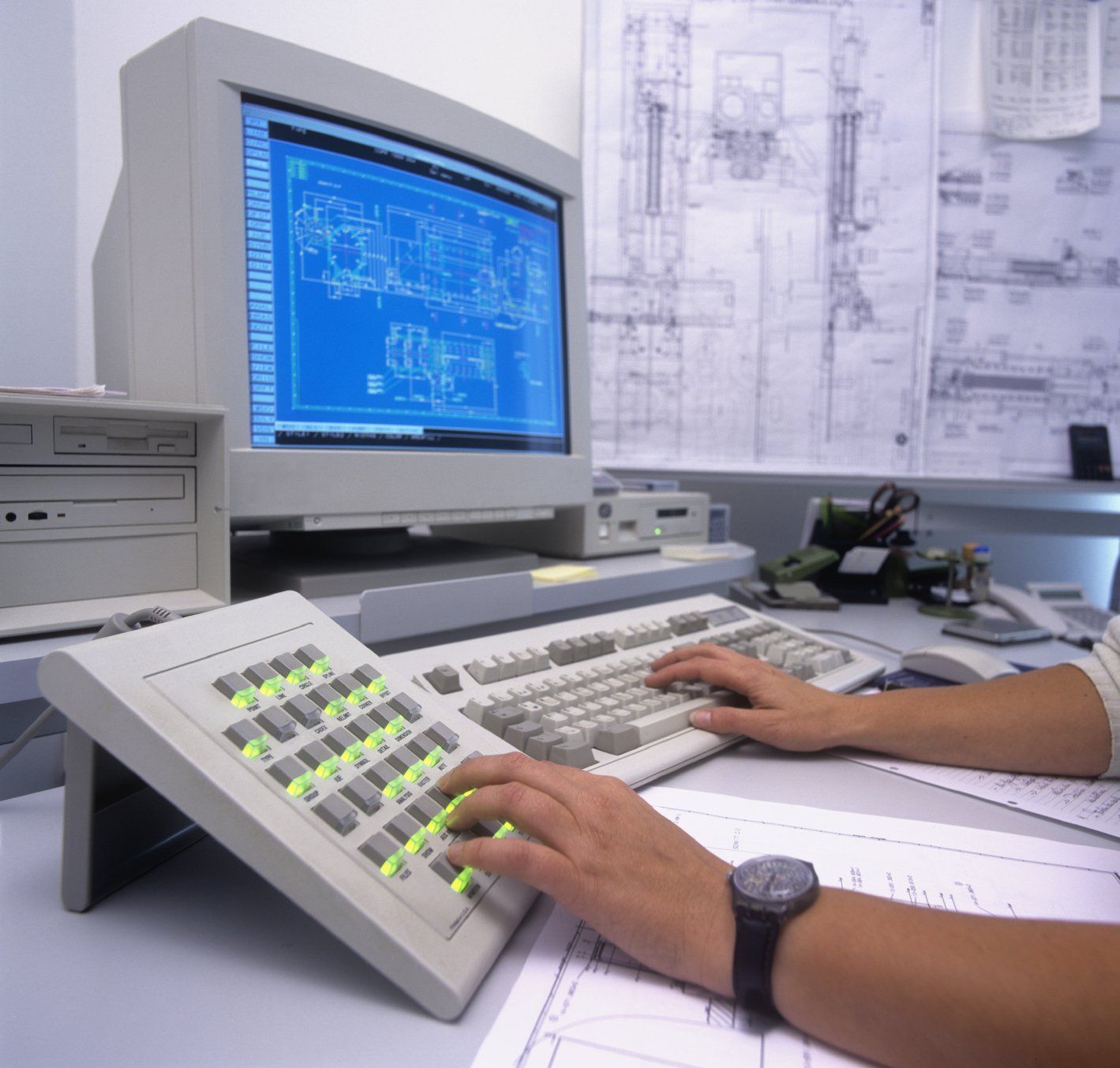 Eine Person bedient einen Computer, eine Tastatur und ein Bedienfeld; im Hintergrund sind Baupläne zu sehen.