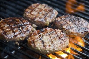 Vier gegrillte Hamburger-Patties auf einem Grill mit Flammen, die Grillstreifen aufweisen.