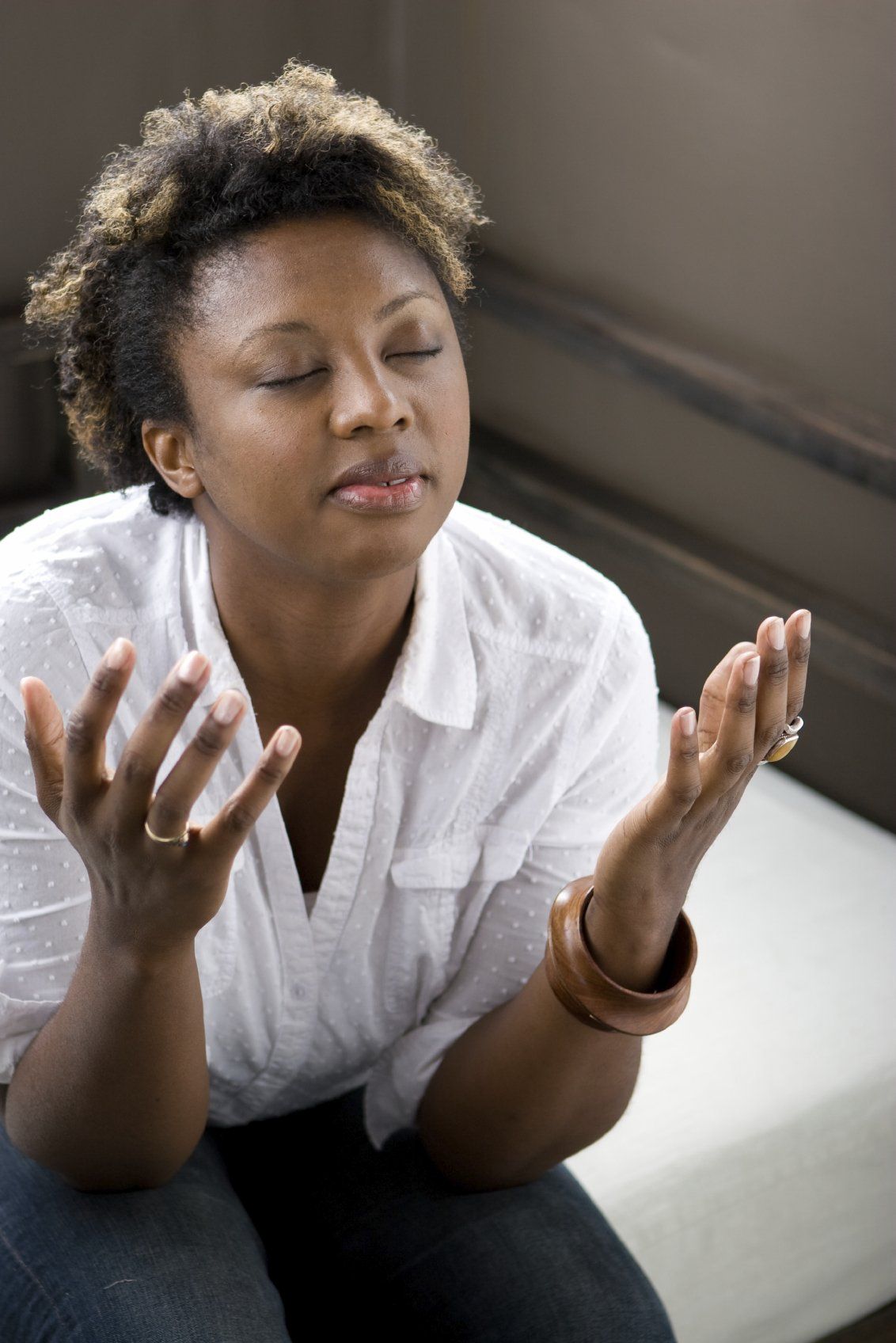 Young black woman, sitting on a bed with eyes closed and hands open in prayer.