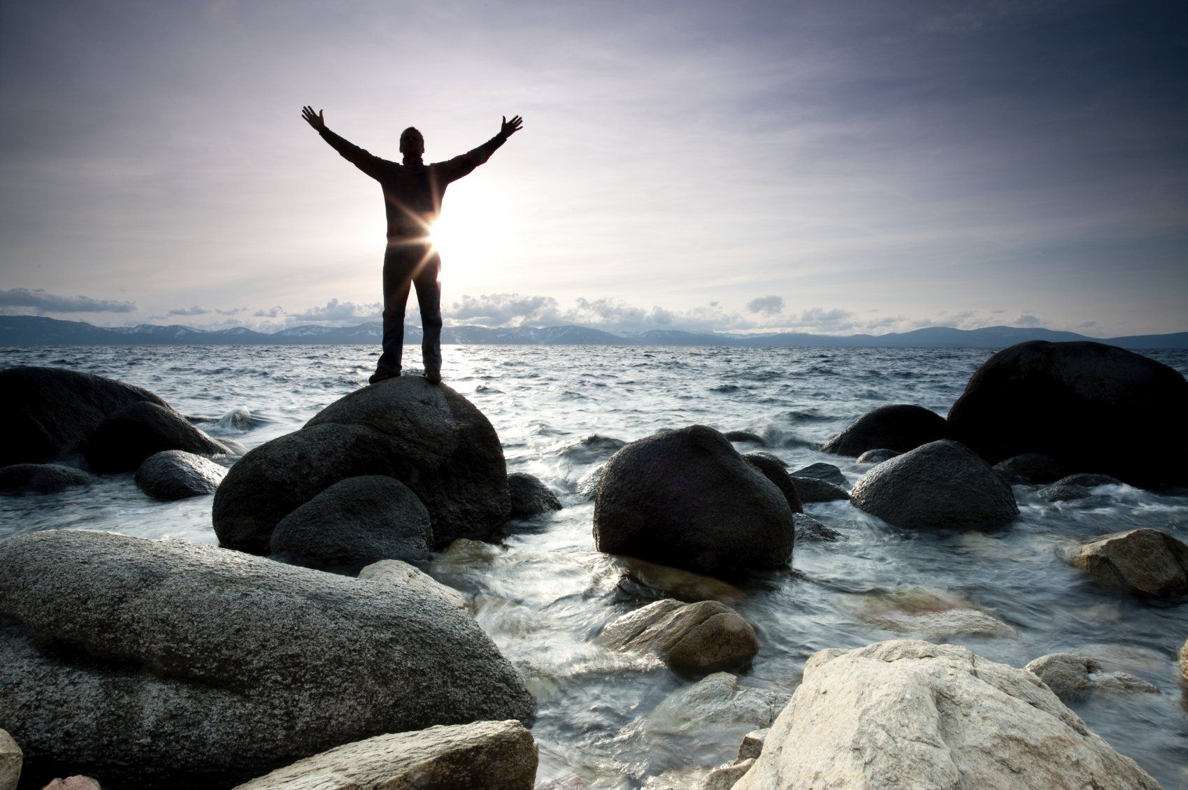 Ein Mann, der die Arme nach oben reiißt dabei auf einen Stein steht, der im Meerwasser liegt. Im Hintergrund eine wunderschöne Berglandschaft