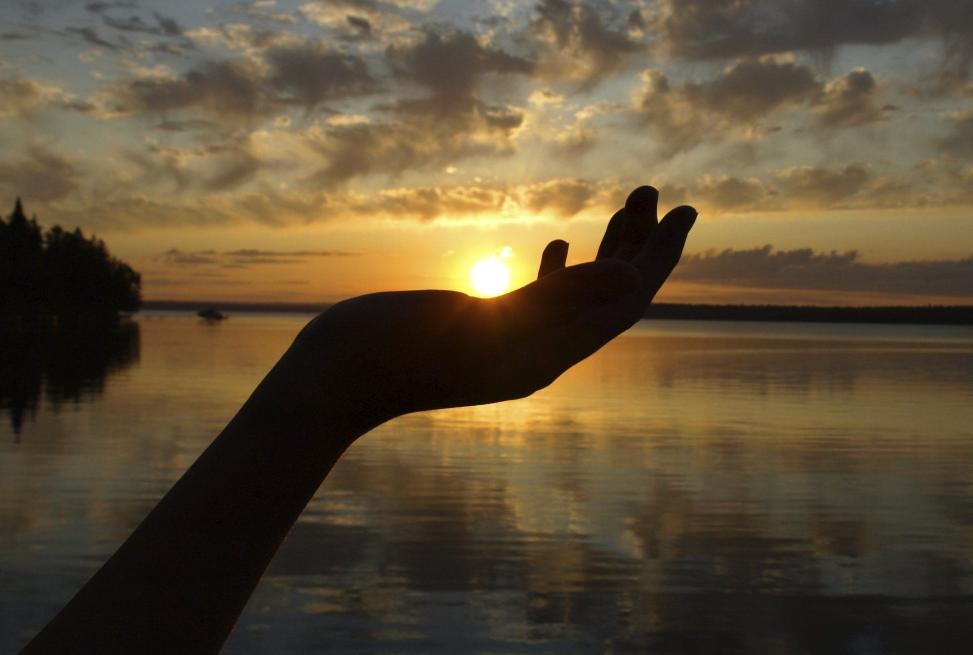 A hand holding the setting sun over reflective water.
