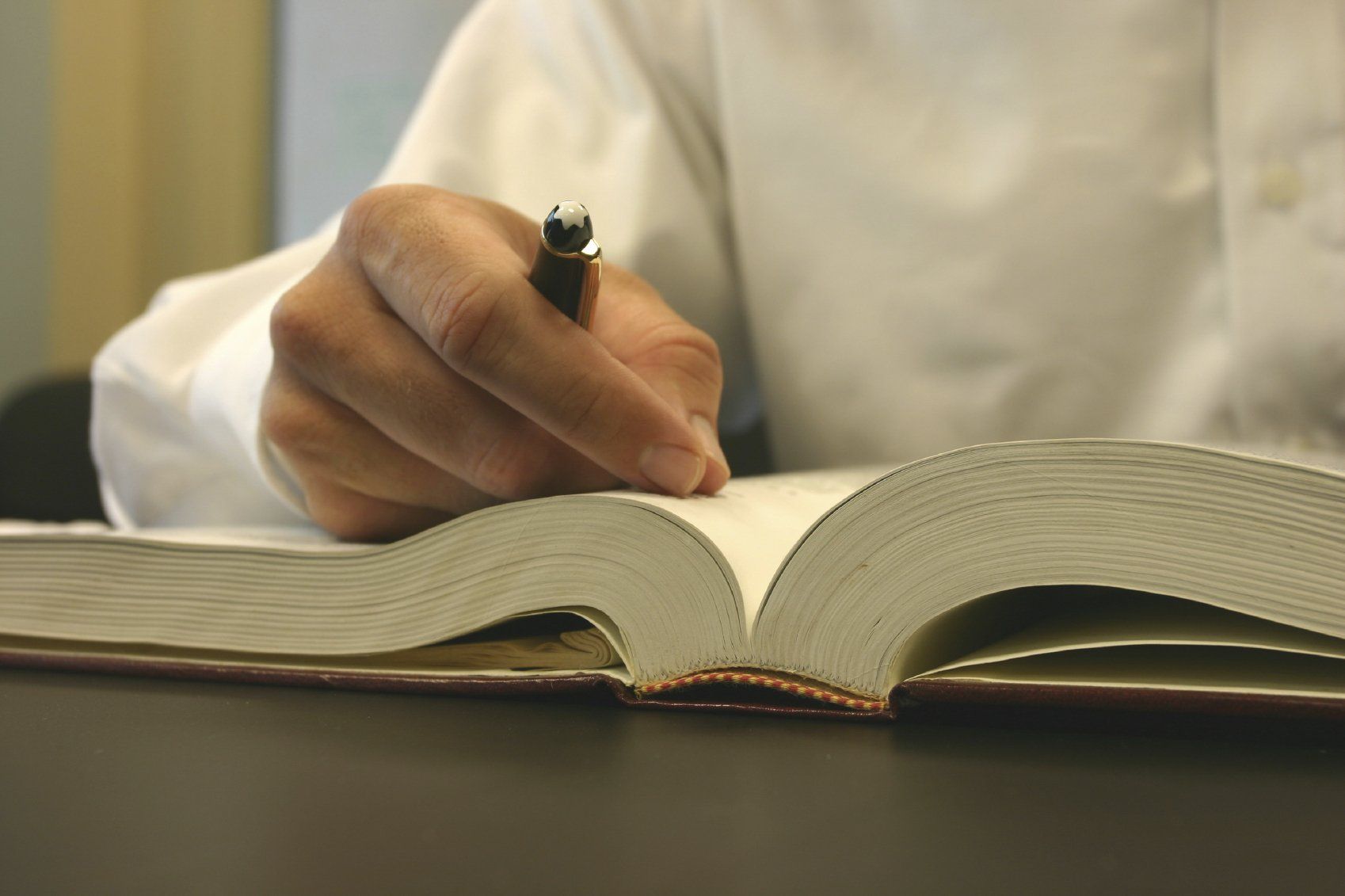 Mann im weißem Hemd und mit Stift in der Hand sitzt an einem Tisch vor geöffnetem Buch