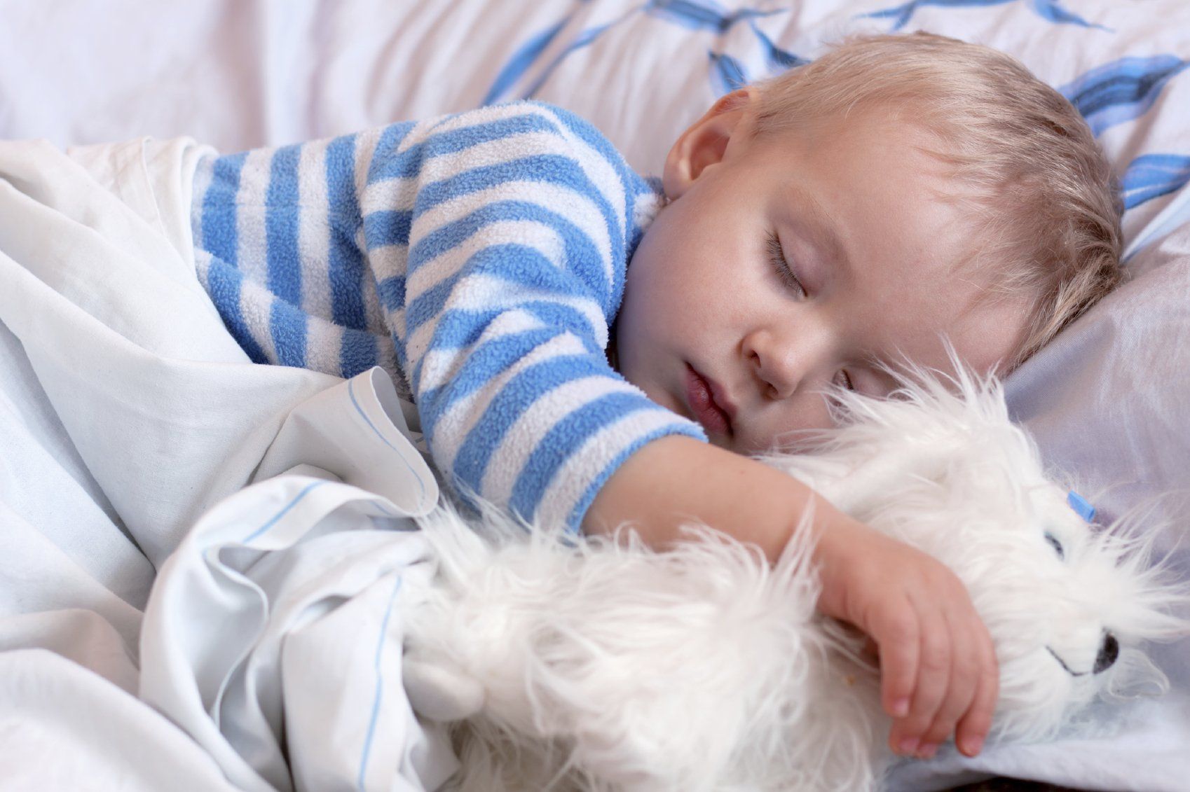 Young child, in striped, blue pyjamas asleep with his white, fluffy dog.