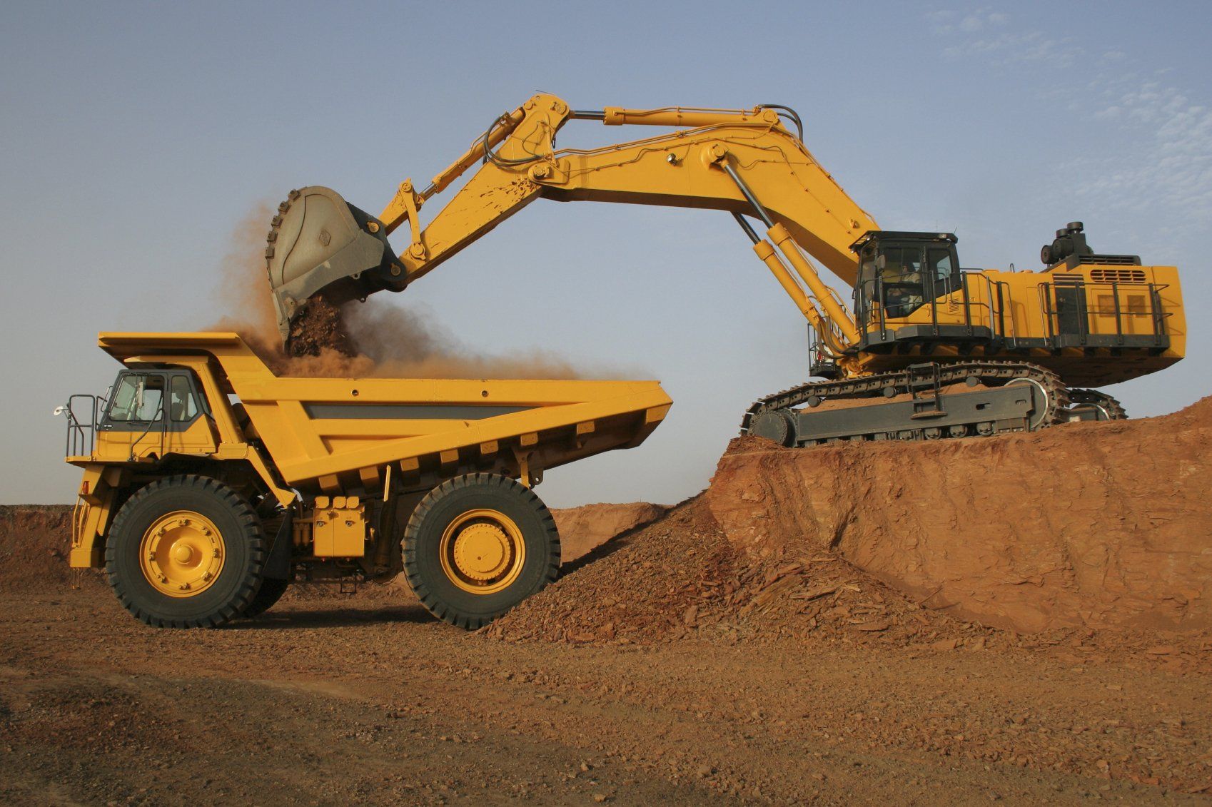 Ein gelber Bagger belädt einen gelben Muldenkipper im Freien mit Rohstoffen.