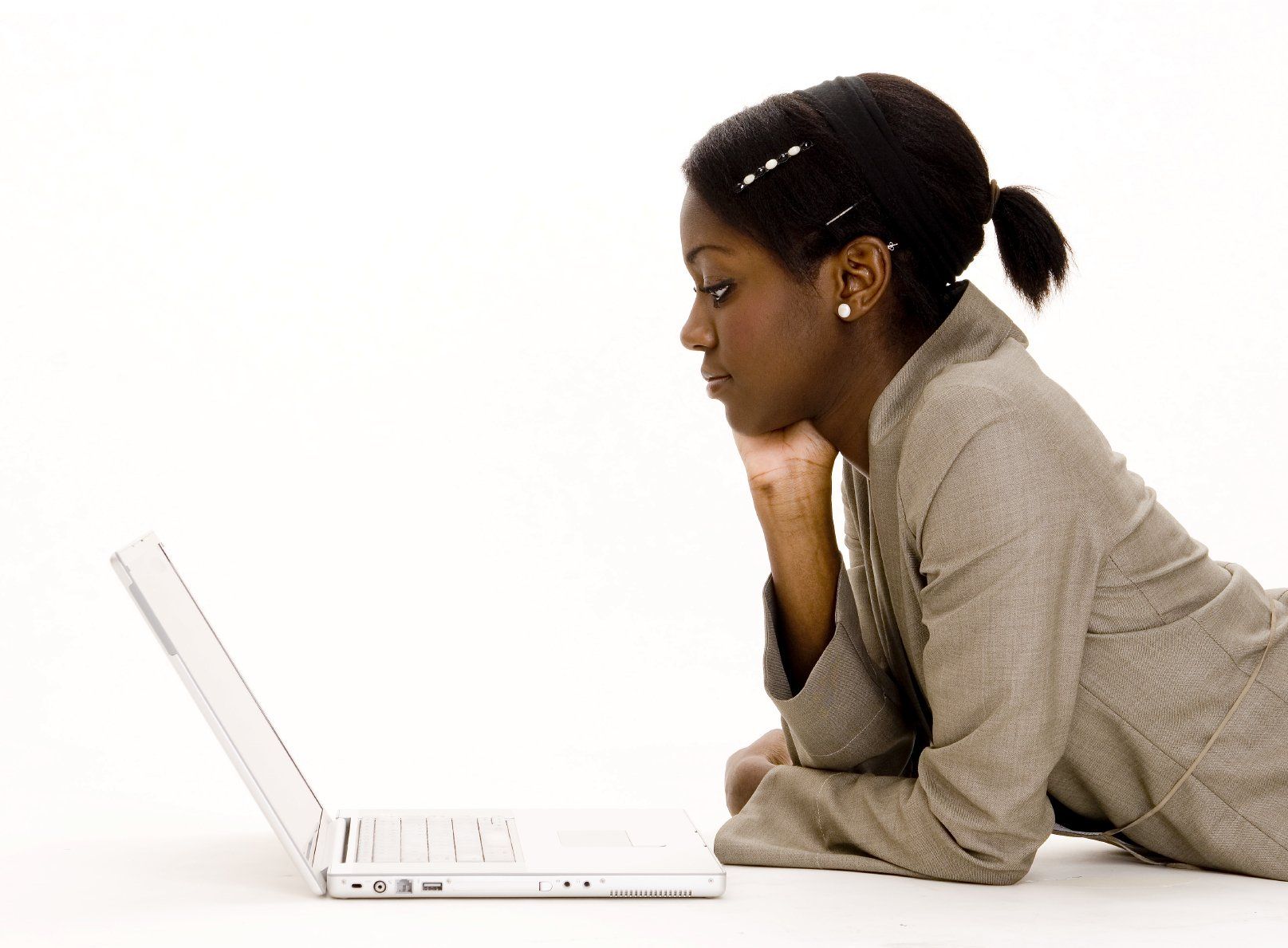 Young, black girl in tan jacket lying on floor with right hand on chin staring at white laptop