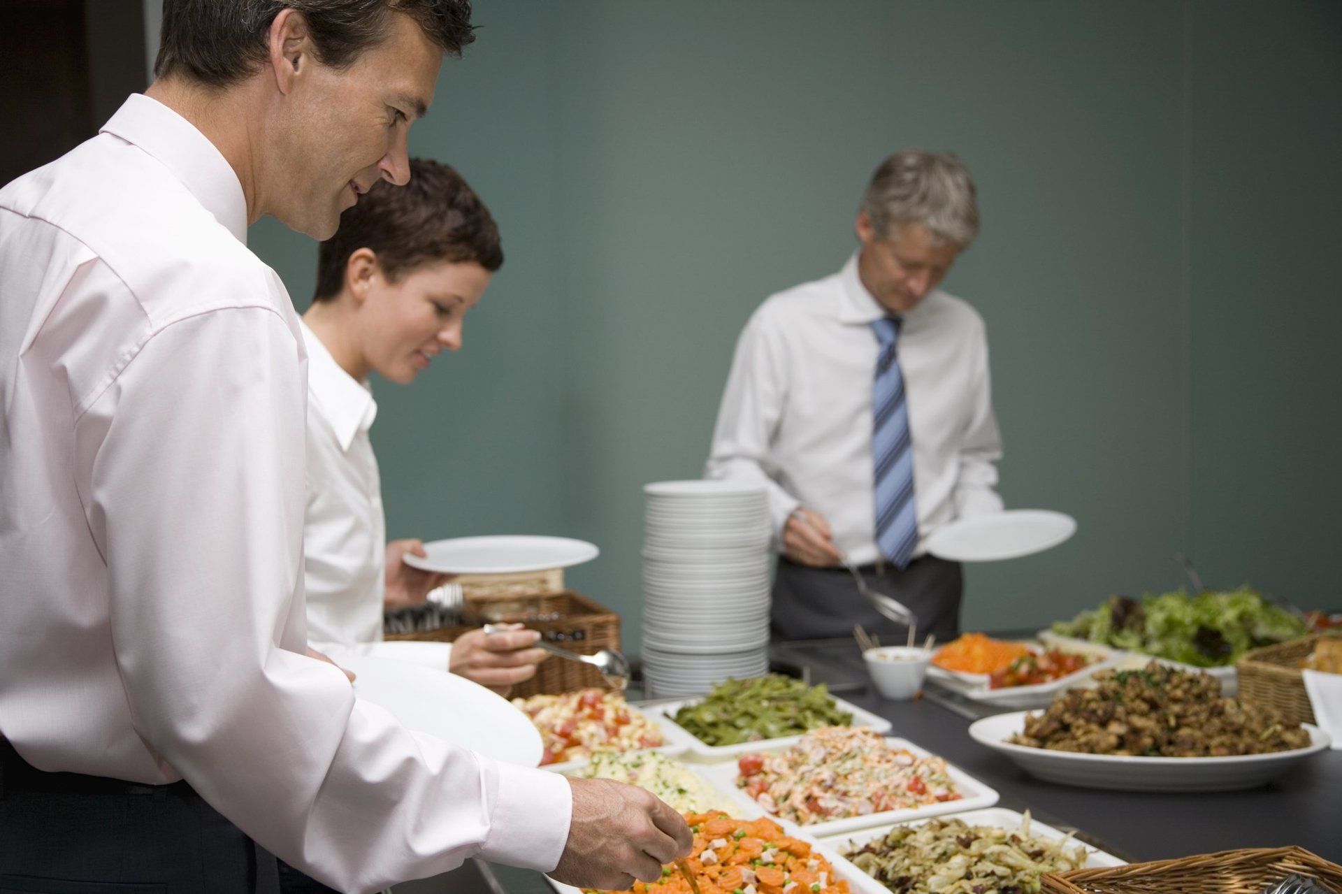 Two men and one woman, dressed in business attire, selecting food from a smorgasbord(buffet)