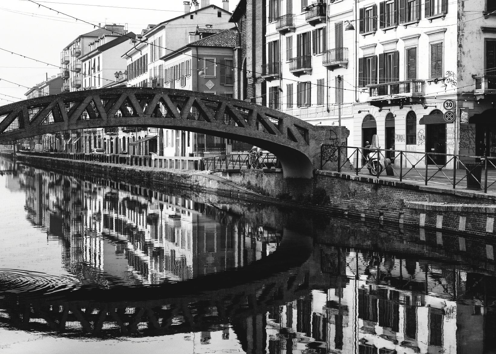 A black and white photo of old buildings on the banks of a river.  An arched bridge spans the river.