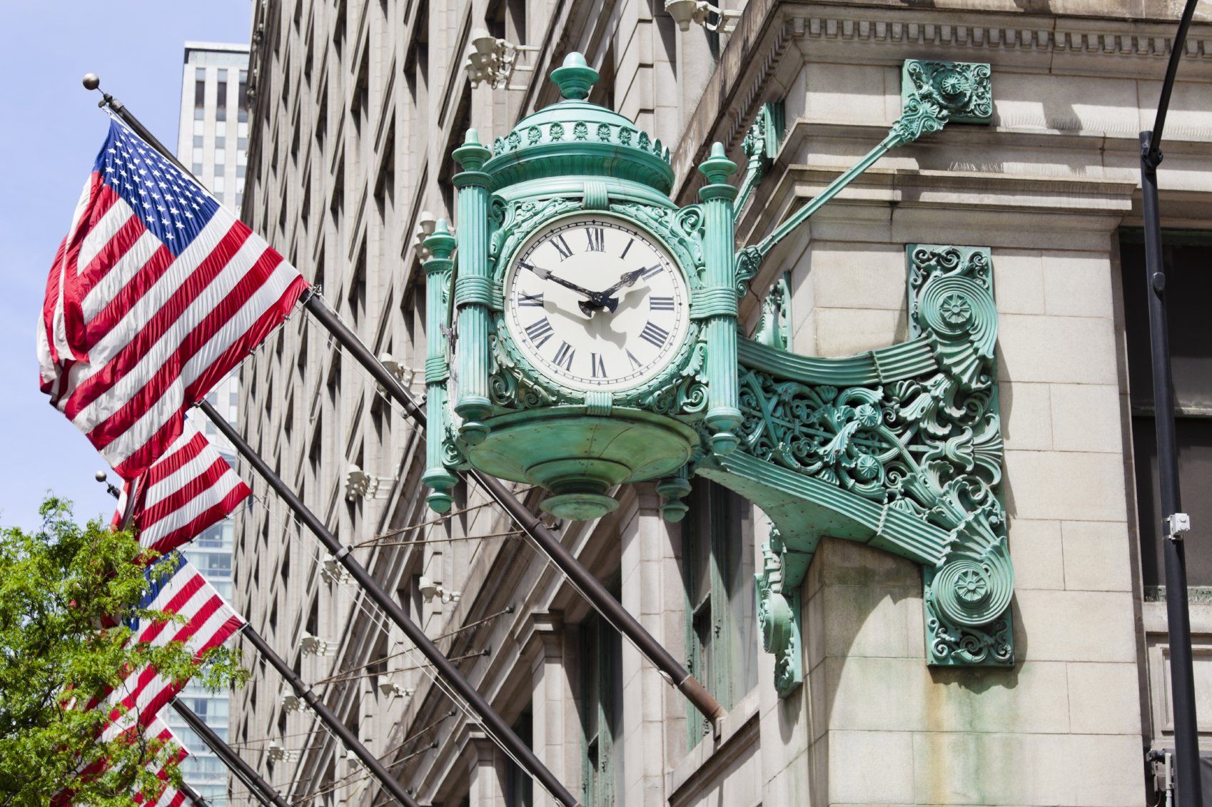 Ancient green clock hung on an old building.  A series of  American flags hang on the building, in the background.