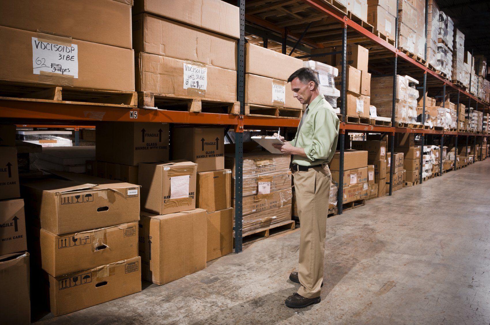 A person in a warehouse with carton boxes, the man is holding something, looking at it and writing on it.