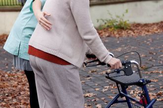 Image shows an elderly person walking using a support frame and a carer with a supporting arm around them