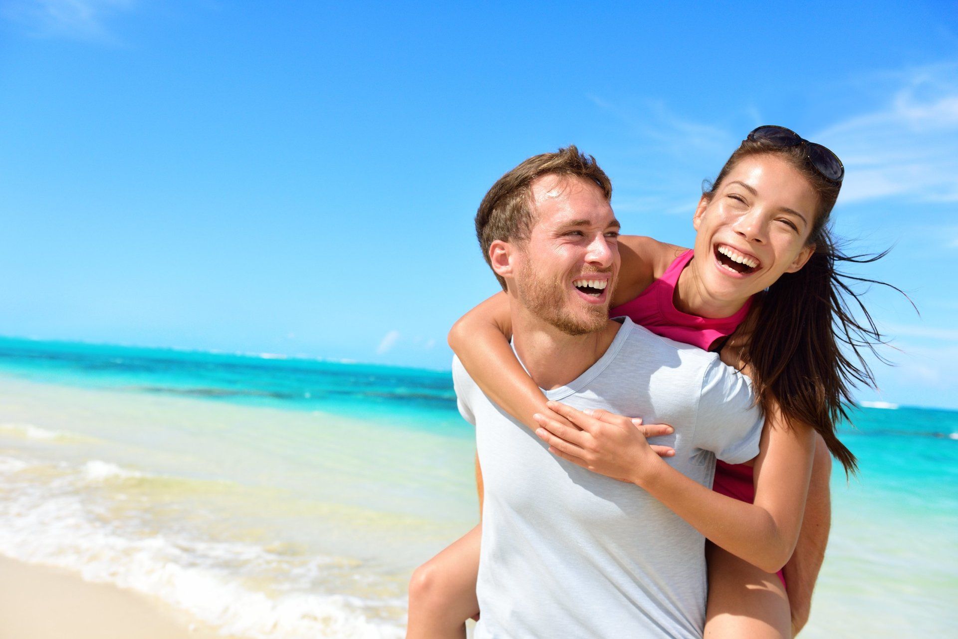 Man in white T shirt  on beach with girlfriend on his back.  Both are laughing.