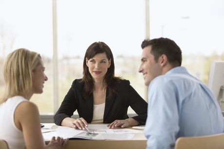 Frau im schwarzen Blazer sitzt mit einem Paar am Tisch; sie besprechen Dokumente. Helles Büroambiente. Vorstellungsgespräch. Kundengespräch. Business.