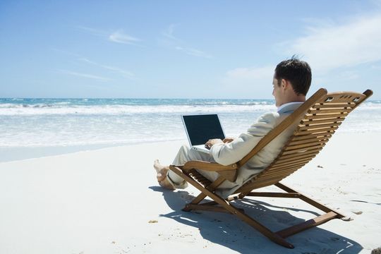 Man in tan suite, no shoes, sitting on a wooden lounge chair on the beach,  and working on his laptop.